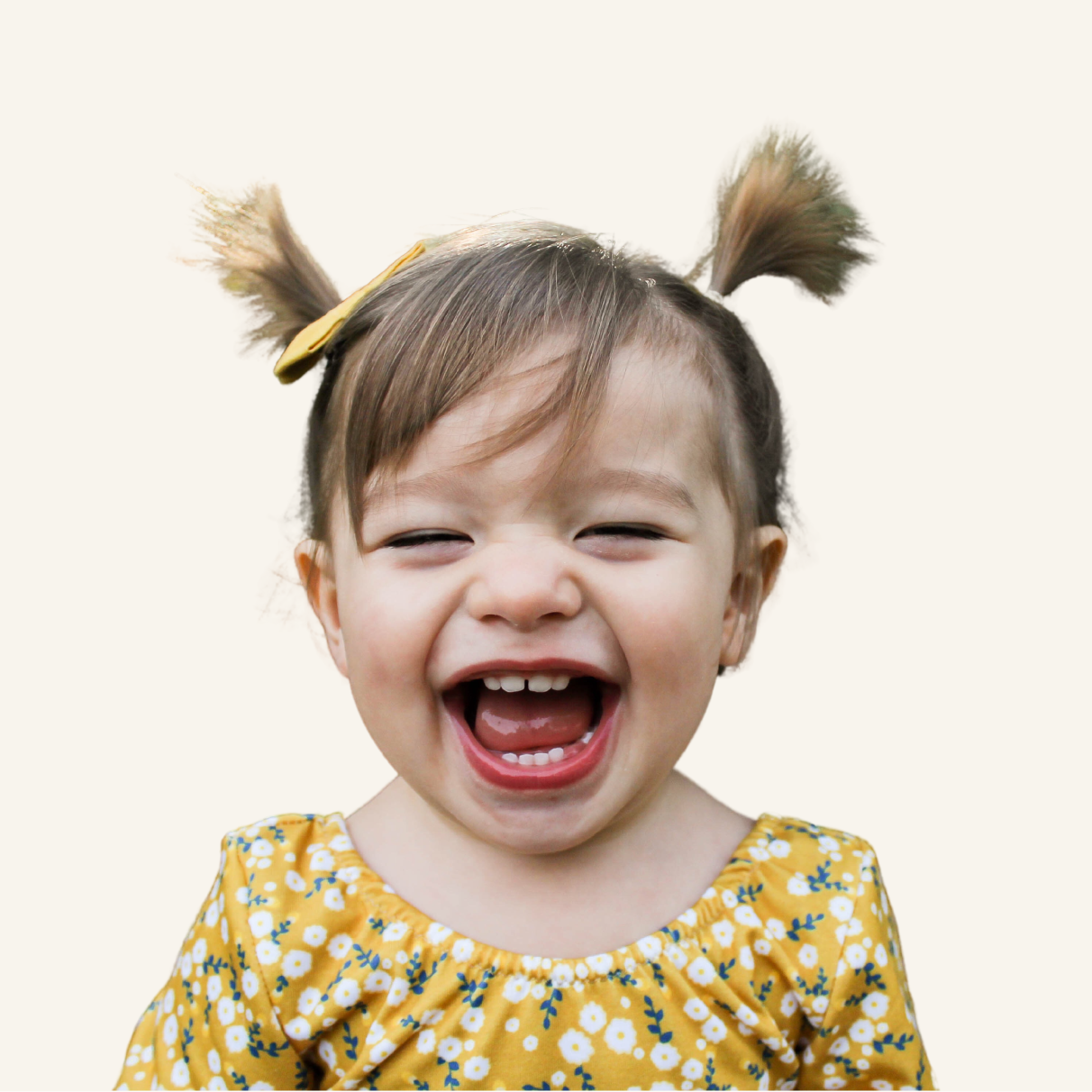A young child with pigtails and a yellow floral dress smiles widely and laughs, facing the camera against a plain light background.