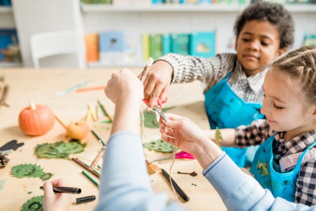 Children at a table work together on an arts and crafts project with leaves, sticks, and pumpkins, wearing blue aprons.