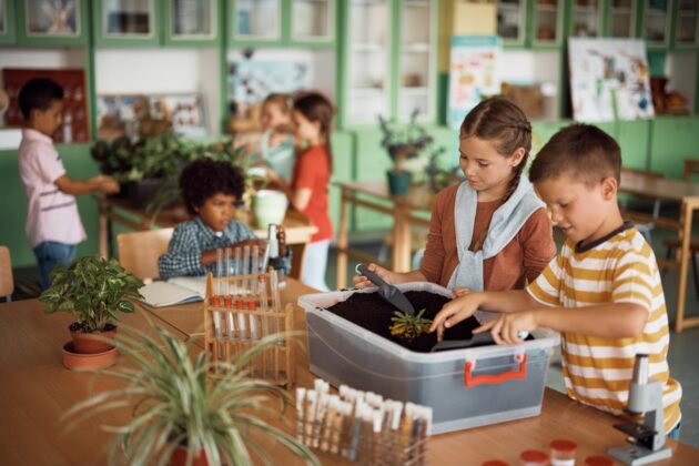 Children in a classroom work in groups on plant science projects, handling soil, plants, and lab equipment on tables, with shelves and posters in the background.