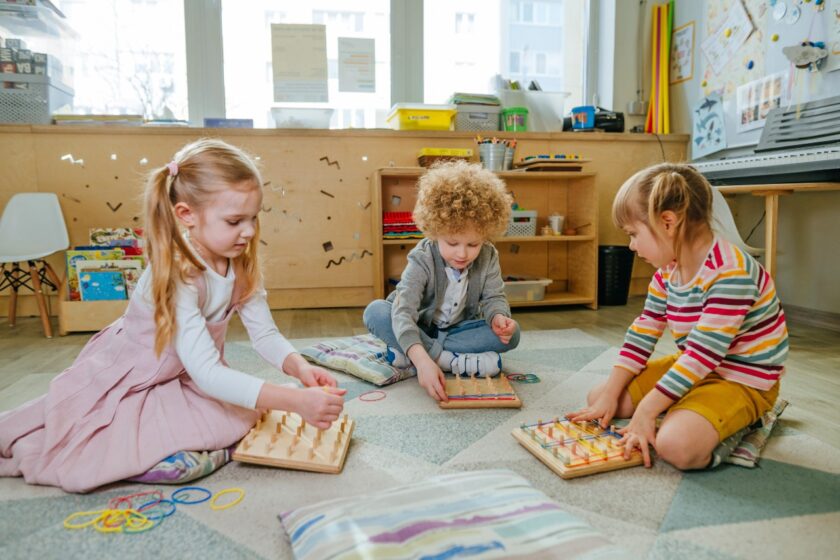 Three young children sit on a carpeted floor in a classroom, playing with wooden peg boards and colorful rubber bands.