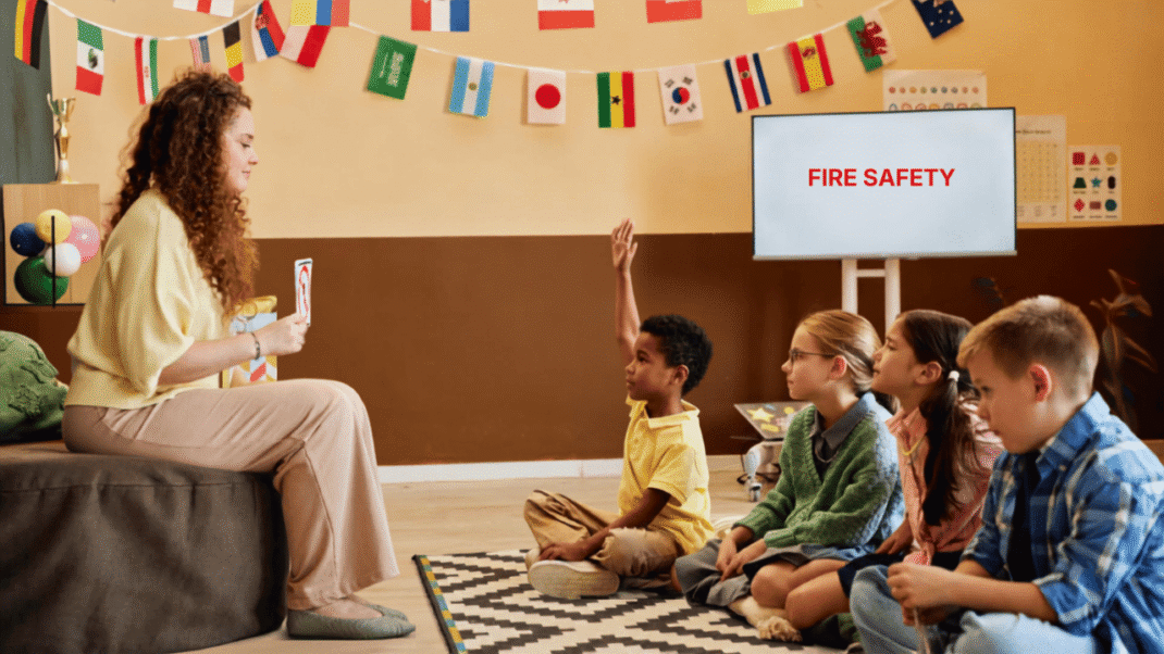 A teacher sits with four children in a classroom. One child raises a hand. A screen displays the words “FIRE SAFETY.” Flags are hanging on the wall in the background.